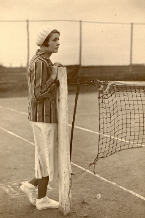 A young woman in tennis clothes from the 1930s era waiting next to the court