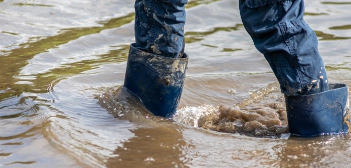 Close up of persons legs walking through dirty flood water, wearing dark blue jeans and black gumboots. Water level coming up over their ankles. 