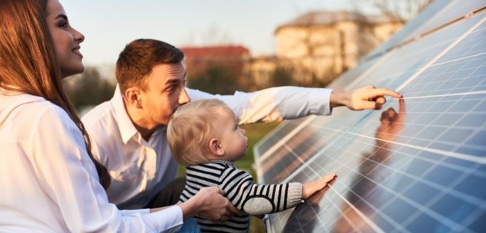 Caucasian straight couple with a blonde baby looking at a solar panel up close. The dad is pointing to the panel, engaging the baby. The mum is helping the baby stand to touch the panel while she smiles looking up to the panel.