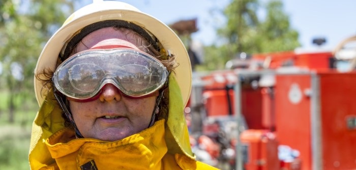 Close up photo of a fire fighter in PPE, feminine presenting with a neutral facial expression standing in front of a fire truck